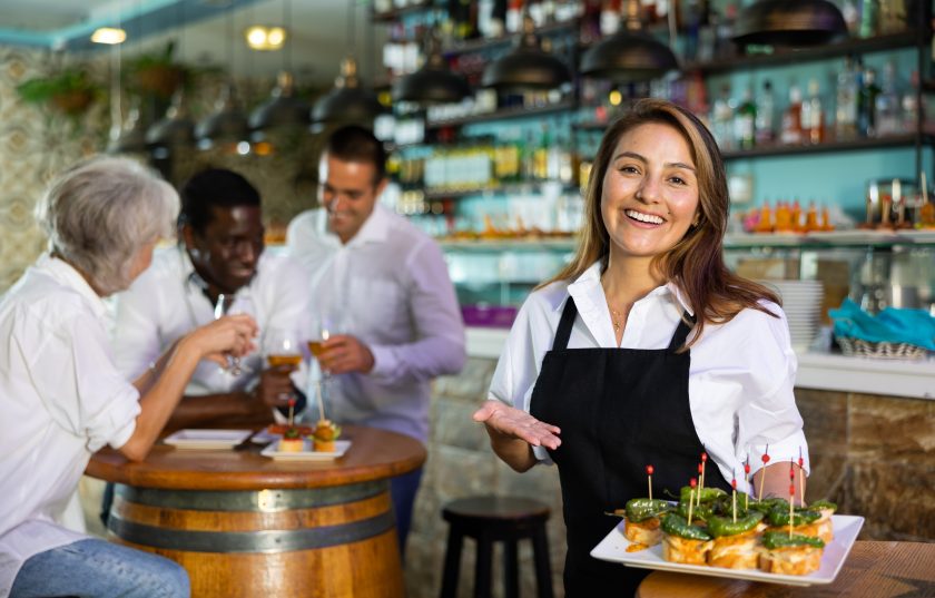Cheerful smiling female waiter holding served tray meeting visitors at bar